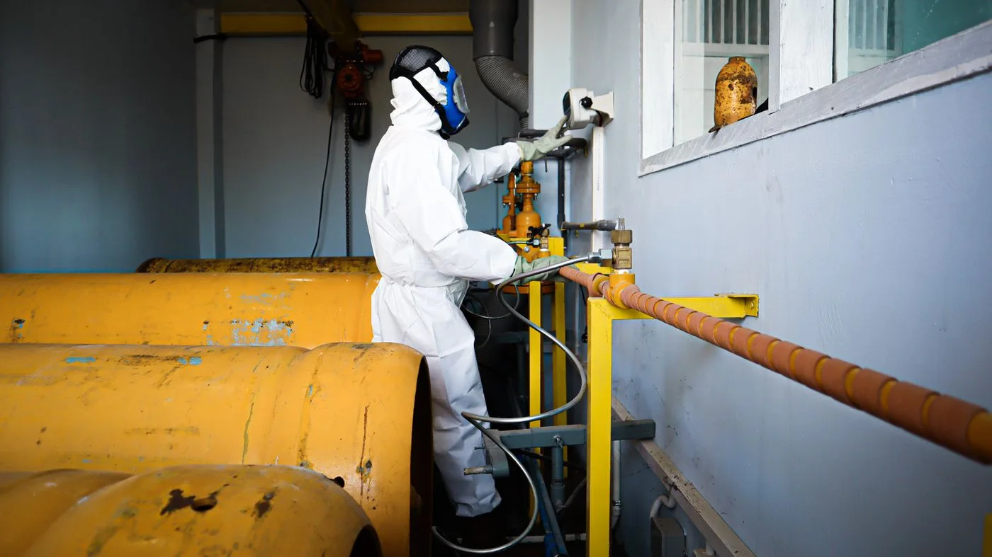 Profissional com traje de proteção e respirador operando válvula próximo a tanques industriais